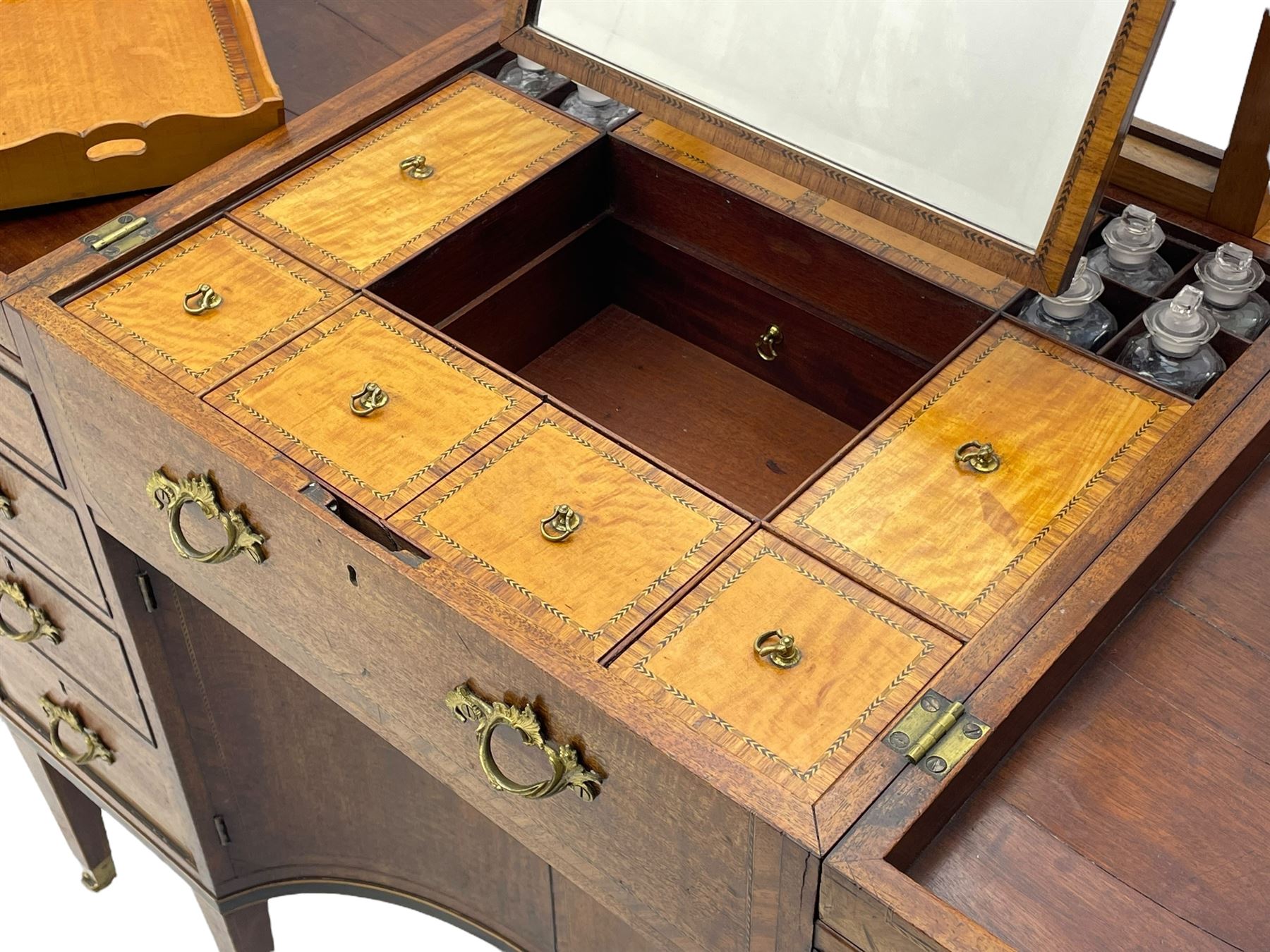 Late 19th century mahogany and satinwood dressing chest, the raised middle section hinges to reveal an interior fitted with lidded compartments, removable tray, single drawer and glass scent bottles, lift-up bevelled mirror with adjustable hinged mechanism, fitted with a further eight cock-beaded graduating drawers and curved kneehole with double cupboard, square tapering supports on brass cups and castors, inlaid throughout with checkered stringing, ornate Rococo cast gilt metal handles