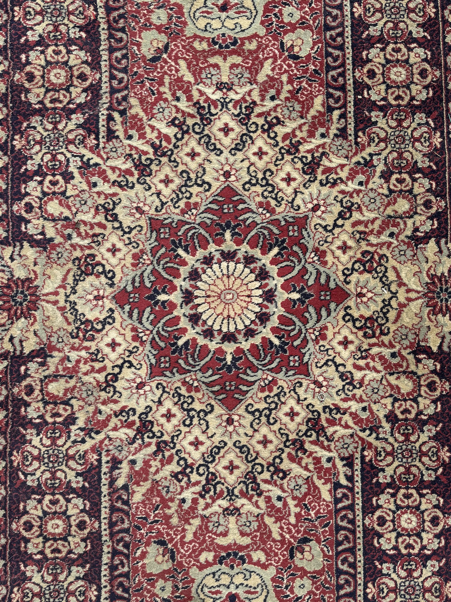 Persian Heriz design red ground carpet, the field decorated with a central star-shaped medallion surrounded by geometric floral motifs and angular vine patterns, the spandrels highlighted with palmette designs, the main border featuring a series of stylised rosettes and leaves against a dark blue ground, enclosed by multiple guard stripes with alternating floral and geometric motifs