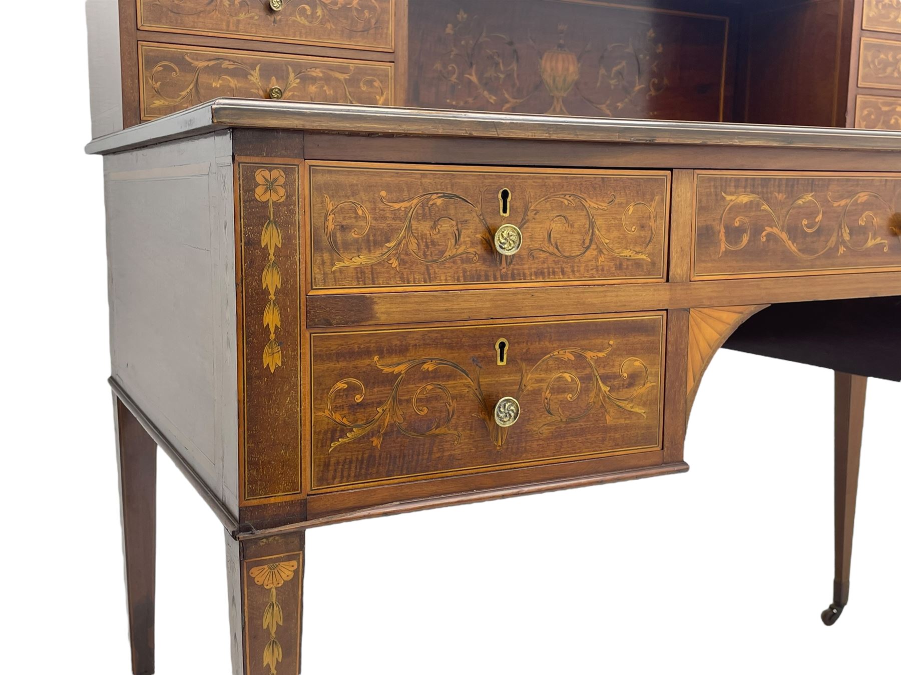 Edwardian inlaid mahogany writing desk, raised galleried back over inlaid central urn with scrolling leafy branches and six small drawers, satinwood banded rectangular moulded top with tooled leather inset, fitted with five drawers, the drawer facias inlaid with scrolling foliage and satinwood banding, square tapering supports inlaid with trailing bell flowers, on brass and ceramic castors 