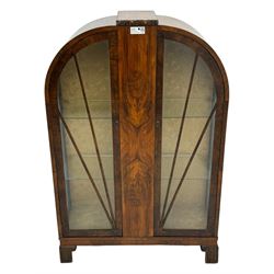 Early 20th century Art Deco walnut display cabinet, with arched top and carved floral crest, central band flanked by two glazed panel doors with decorative mullions, enclosing two glass shelves, on scroll carved bracket feet